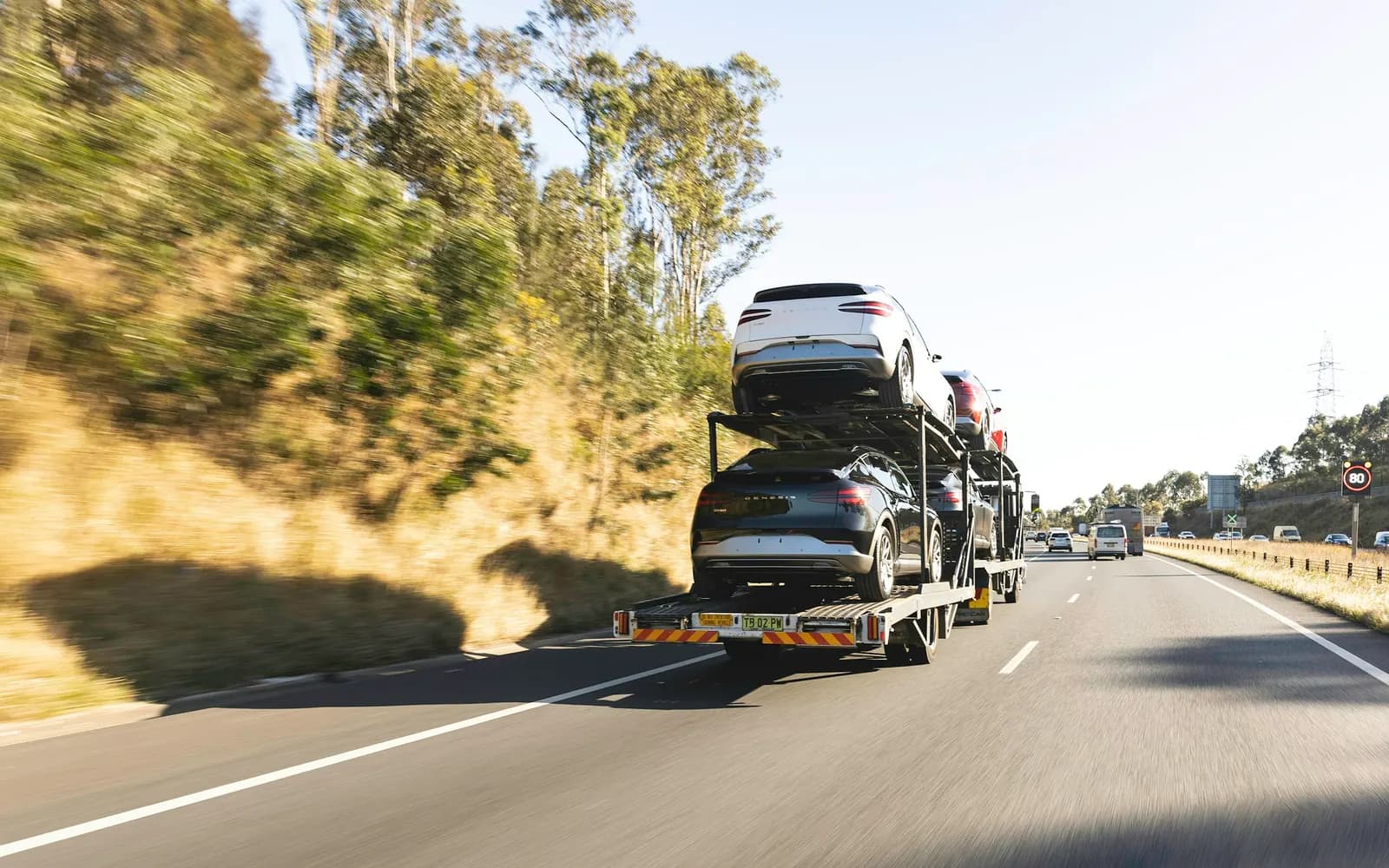 A car transporter carrying a double deck of vehicles along a highway, seen from the lane behind it.