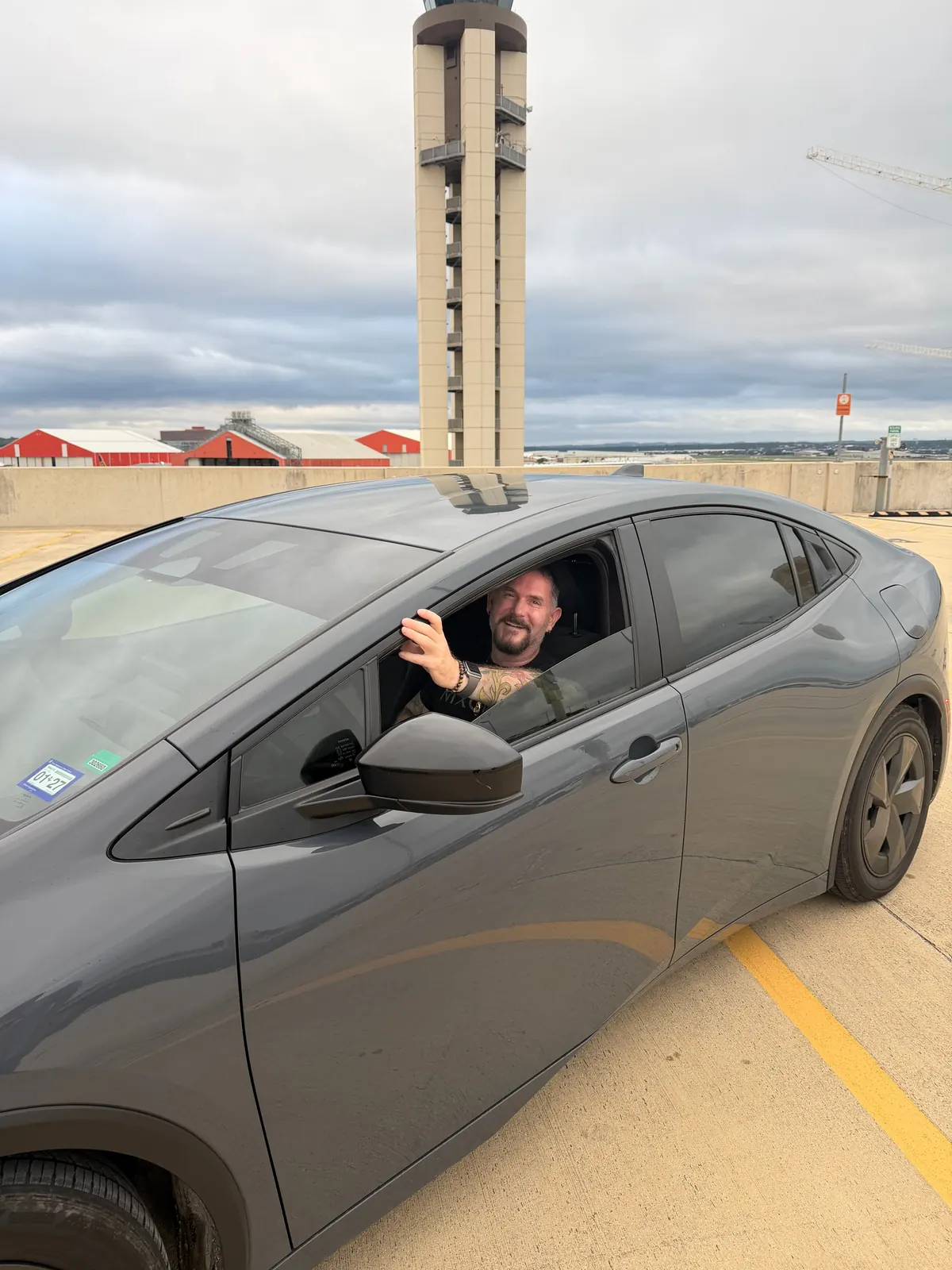 A smiling man leaning out of the open driver’s window of a grey Toyota Prius parked on the top deck of a parking garage, with an airport control tower behind.