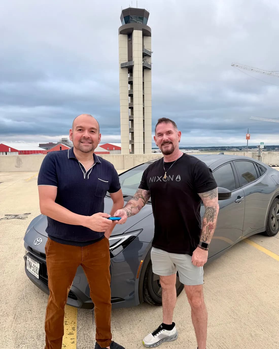 Two men passing a car key between them in front of a grey Toyota Prius on the open top deck of a parking garage, with an airport control tower behind.